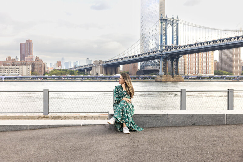 Vanessa Vancour sits on a bench in front of a bridge in New York City over a river. She is wearing a flowy green dress and looking to the right.
