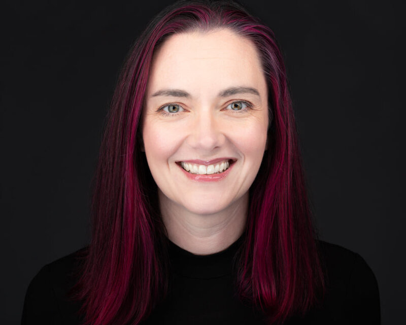 Fort Mill Portrait Photographer Alicia Insley Smith smiling facing the camera wearing black with dark background, woman has pink hair, daark eyebrows and green hazel eyes close up, friendly, joyful  headshot