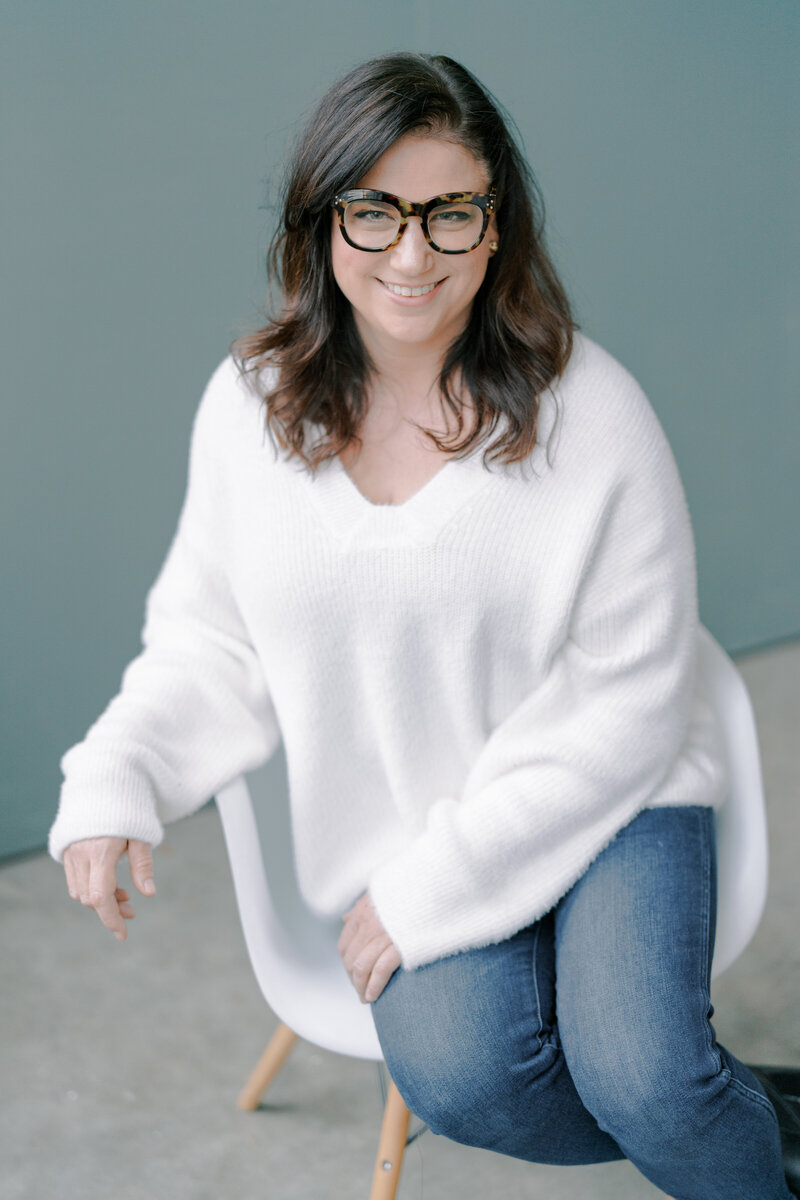 woman in a white fuzzy sweater sitting in a white chair with dark rimmed glasses on smiling at the camera