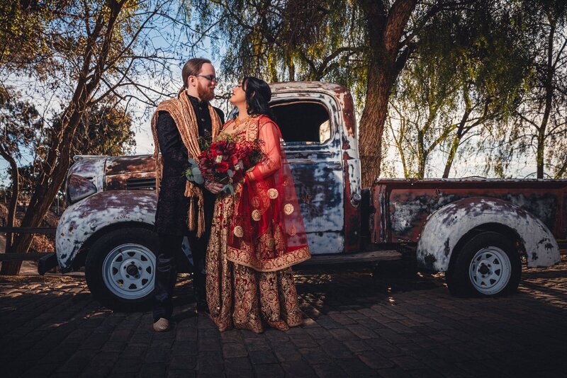 Bride and Groom in Indian Wedding attire embracing with a beautiful red bouquet in front of a rustic truck at Lake Oak Meadows in Temecula.