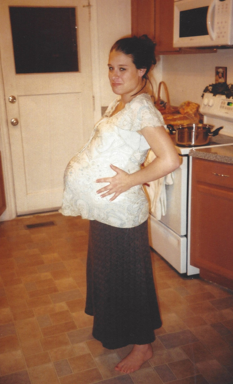 A pregnant person standing in a kitchen, smiling and cradling their belly, captured by Luke and Ashley Photography.