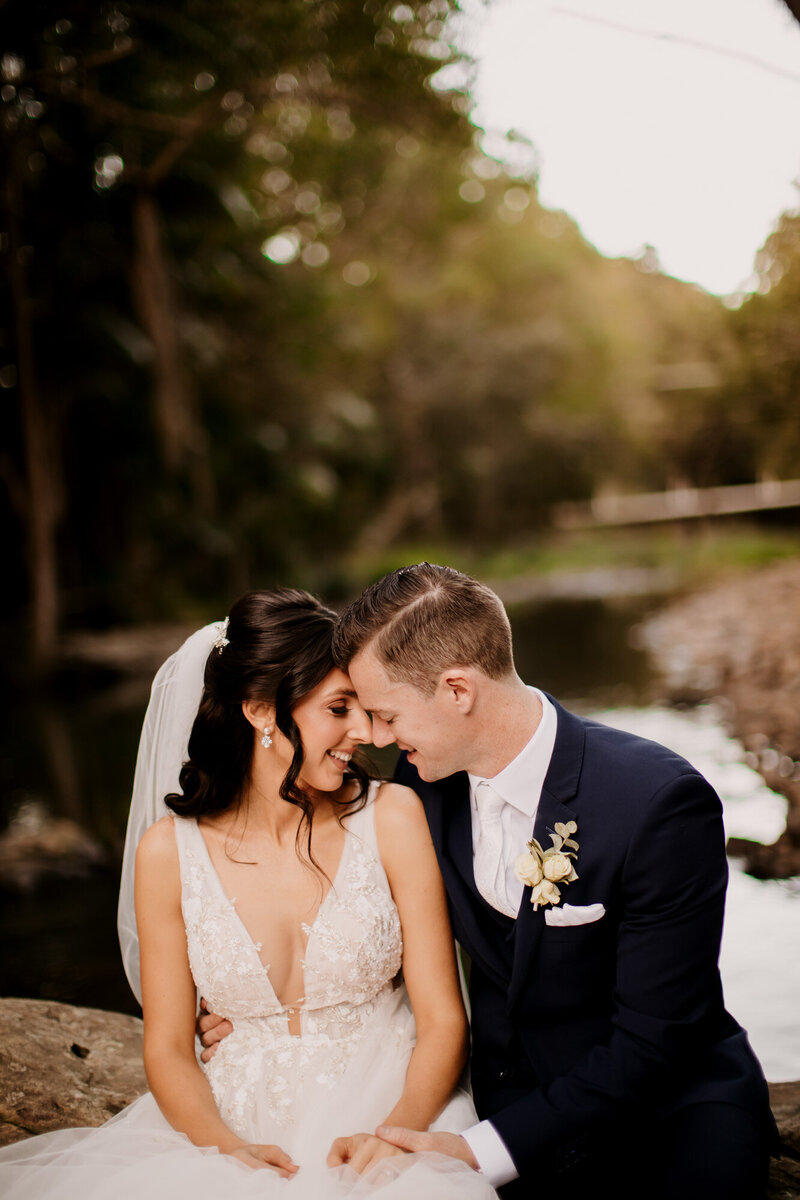 Wedding couple leaning their heads towards each other while sitting on a rock