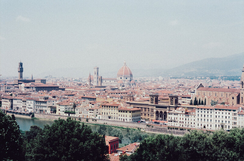A hazy panoramic view of Florence with the river in the foreground, rooftops stretching across the scene, and the iconic dome rising in the distance.