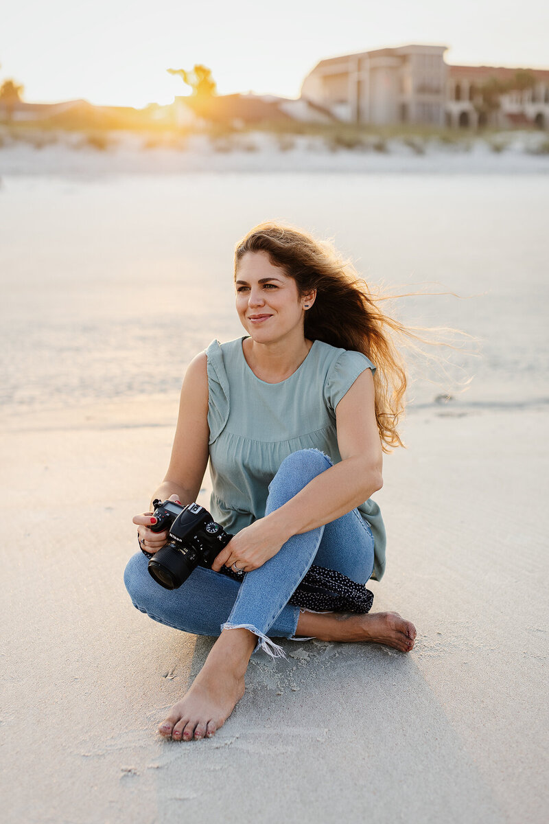 Maternity photographer for Erin Elyse Photography in jeans sitting on beach with camera in Atlantic Beach, FL.