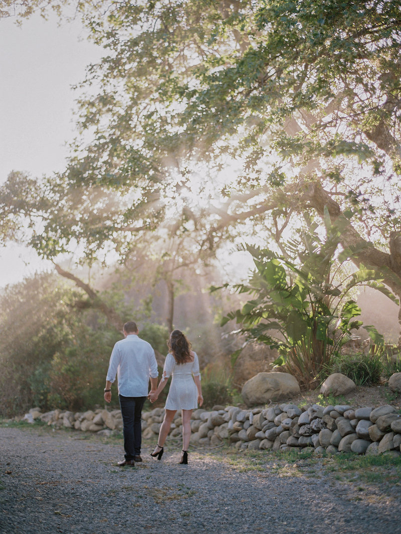a couple walking towards the beach on a road with trees. the light is shining through the leaves in a beautiful way