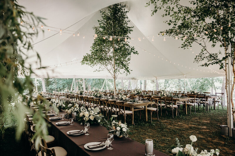 A tented wedding featuring long wood harvest tables and trees planted inside the tent at a private property wedding in Cobden Ontario