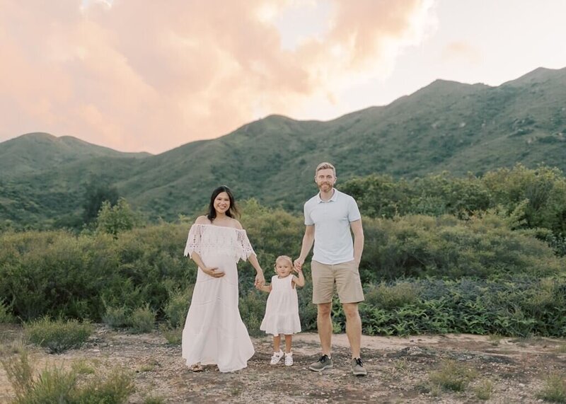 Parents holding their toddler's hand against a grassy mountain backdrop.