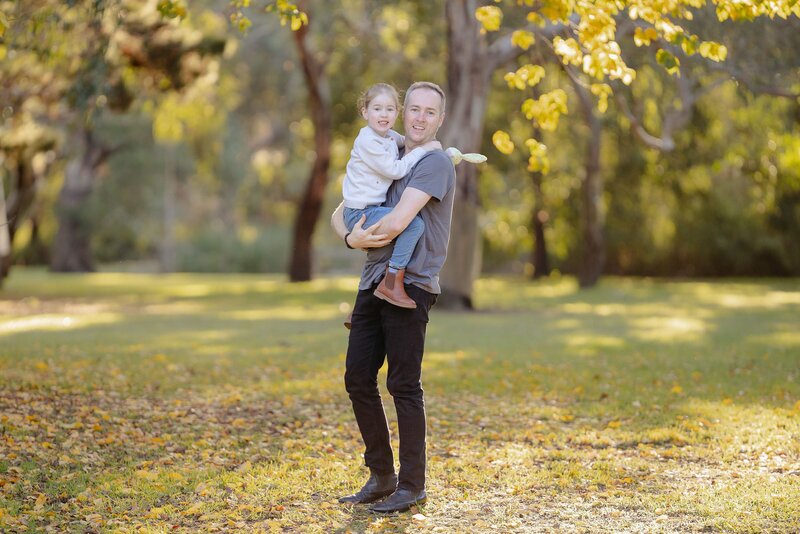 Father and daughter portrait in Geelong captured in soft golden light, with dad holding his smiling daughter in a peaceful park surrounded by autumn trees.
