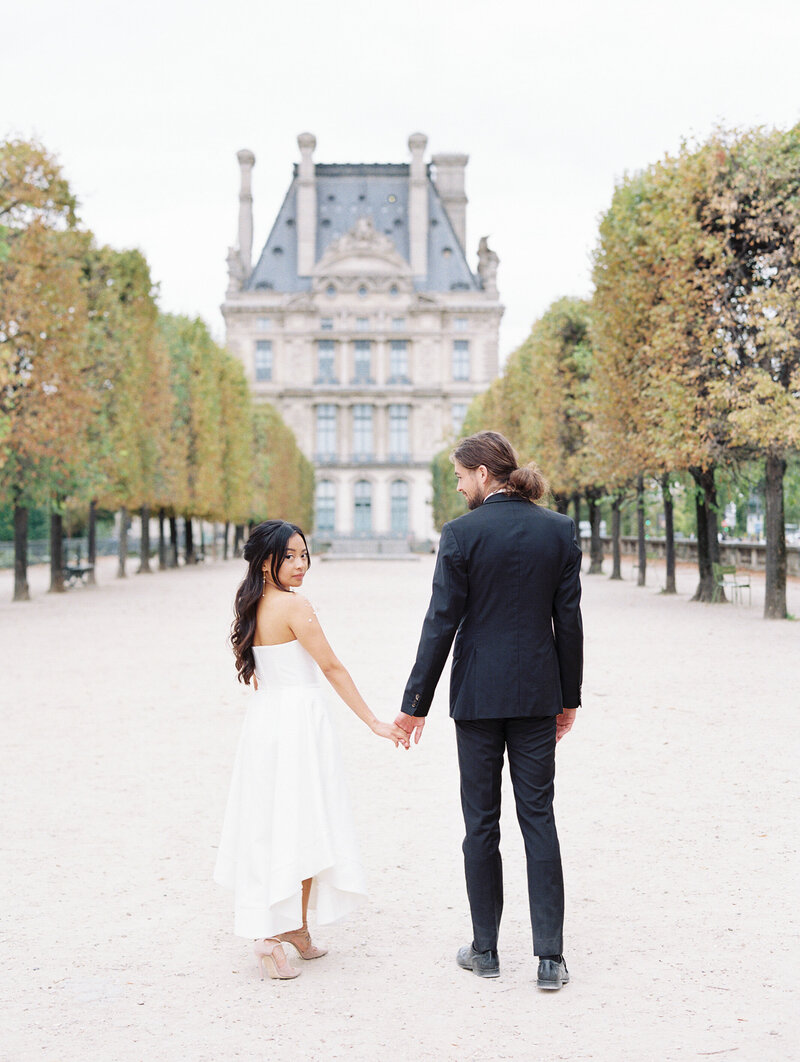 A posed engagement photo of a man and women with their backs facing the camera looking back over their shoulders. They are outside on a large light stone walkway leading to a historic Parisian building. Short round tress line the pathway. The woman where a white strapless dress, and the man wears a black suite.