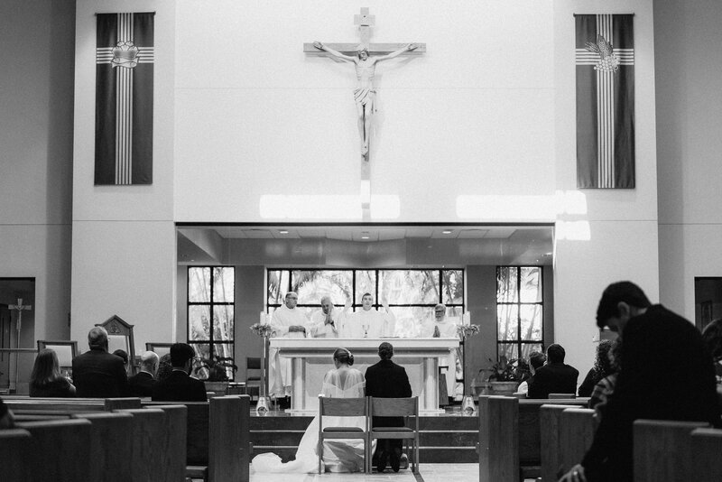 Black and white photo taken from the back of the church of the couple sitting at the altar in a Catholic church wedding. Photo taken by Orlando Wedding Photographer Four Loves Photos and Film.