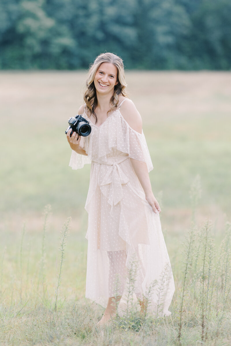A headshot of a family, newborn photographer in a white dress in a field with her camera