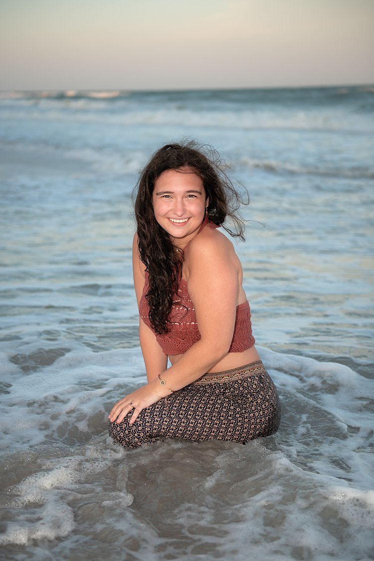 High school senior girl in harem pants and crocheted crop top kneeling in water as the ocean swirls around her