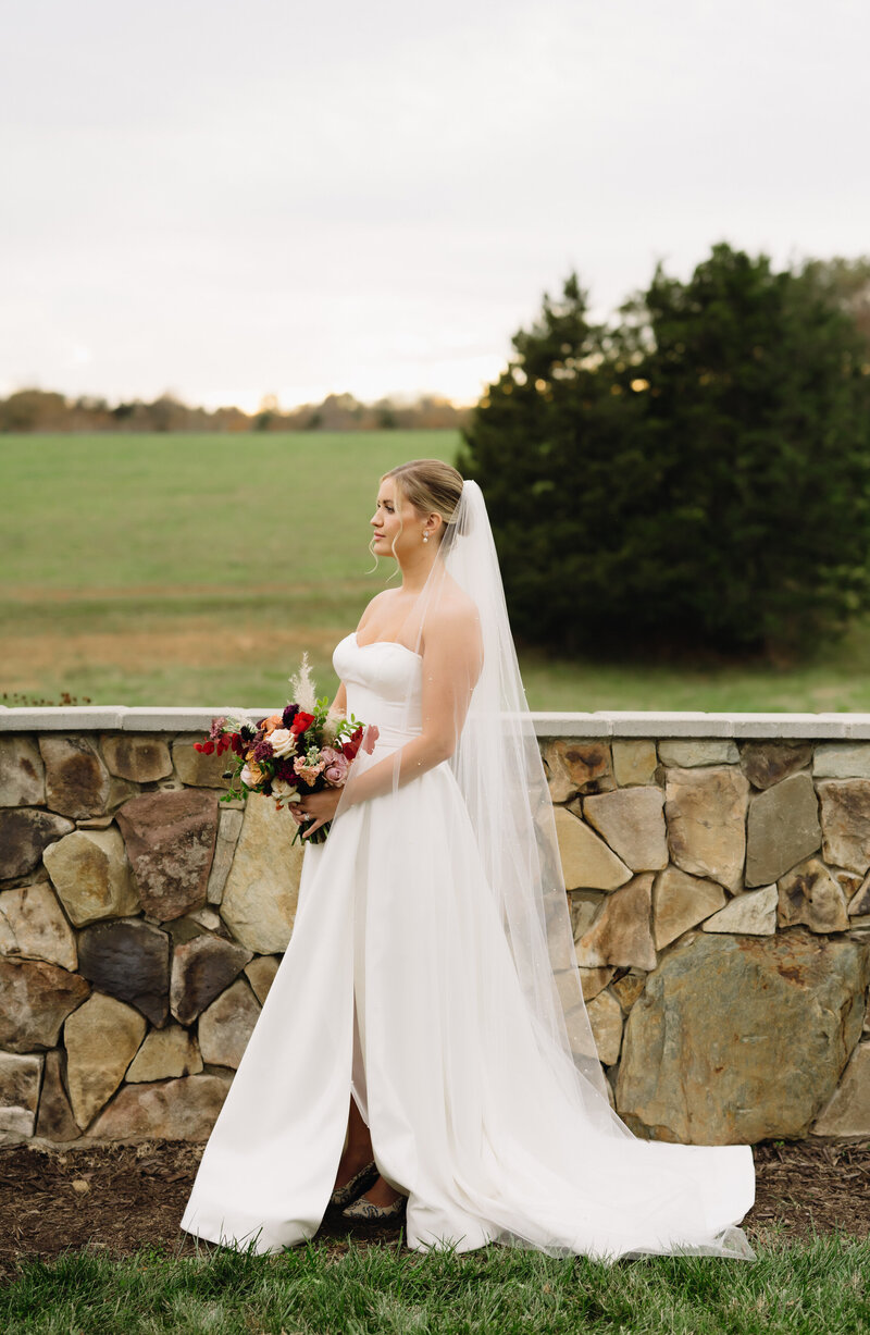 bride standing next to a stone half wall at a luxury northern virginia wedding venue holding a bouquet at her hips and looking off into the distance