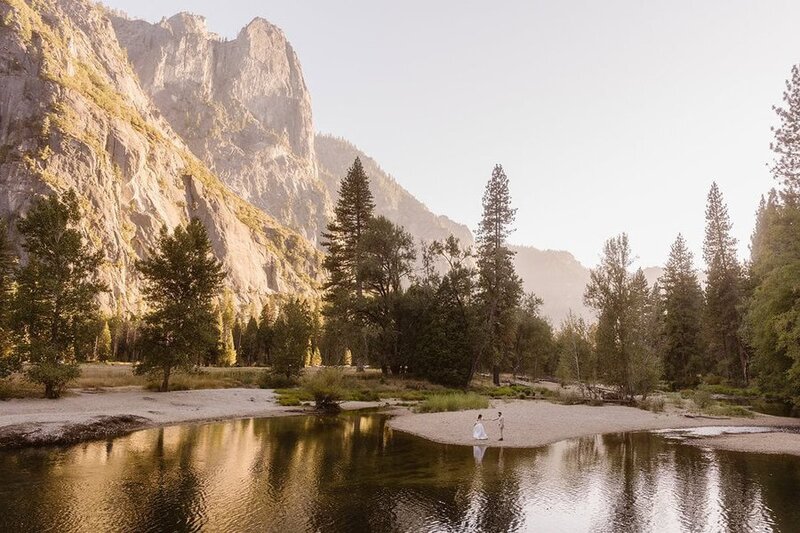 A couple walks hand-in-hand on a small sandbar surrounded by water in a forested valley, with tall cliffs and pine trees lit by soft morning or evening light in Yosemite National Park.