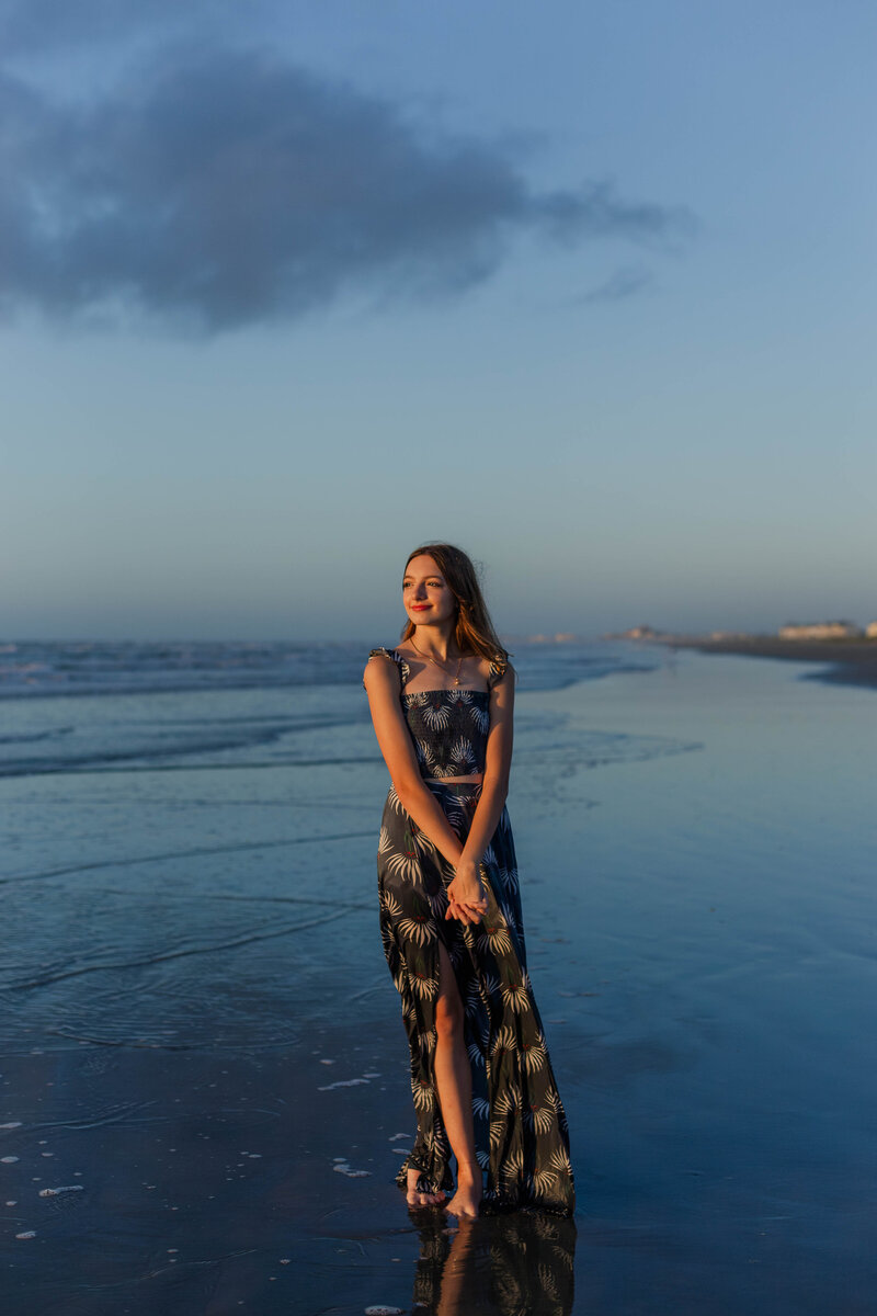sixteen year old girl stands in a dark blue dress while holding her hands and watching the sunrise over Galveston Island