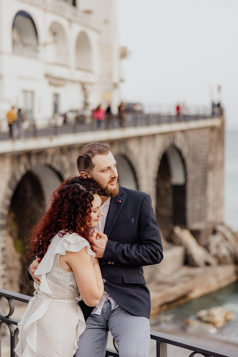 A couple embracing near a stone bridge by the water, with the groom gently holding the bride as she leans into him, captured during a romantic coastal engagement session by a UK wedding photographer.