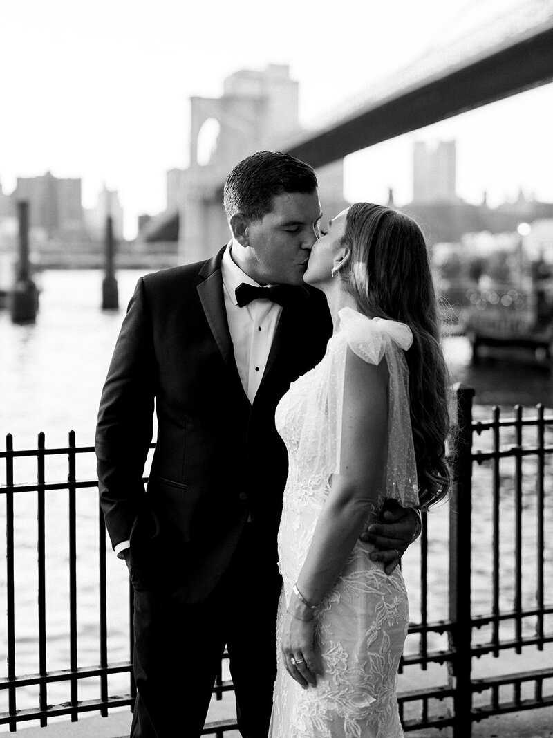 Bride and groom kissing with the Brooklyn Bridge in the background, black and white, groom's arm is around the brides waist.