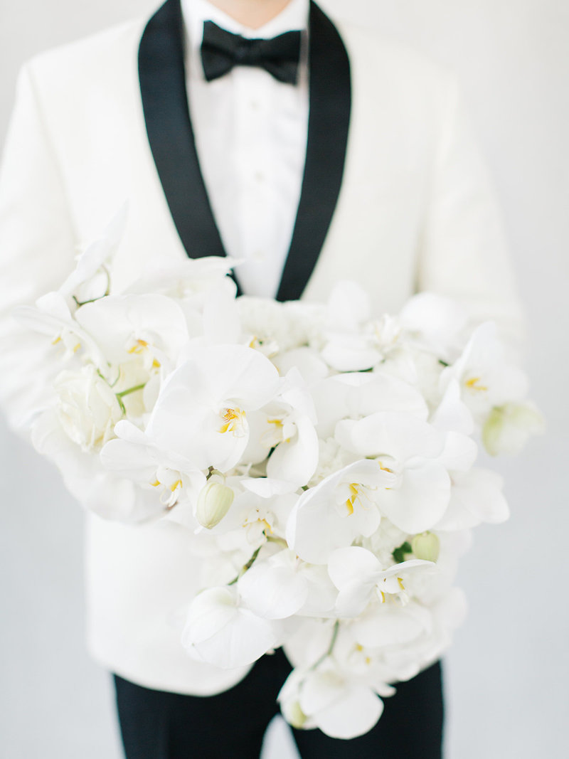 This groom is wearing a classic black and white dinner jacket holding an all white orchid bouquet