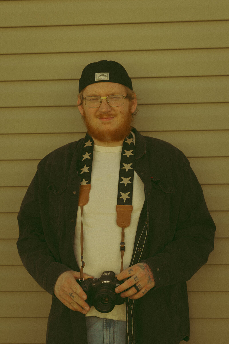 a man smiles into the camera as he holds a camera in his tattoo covered hands
