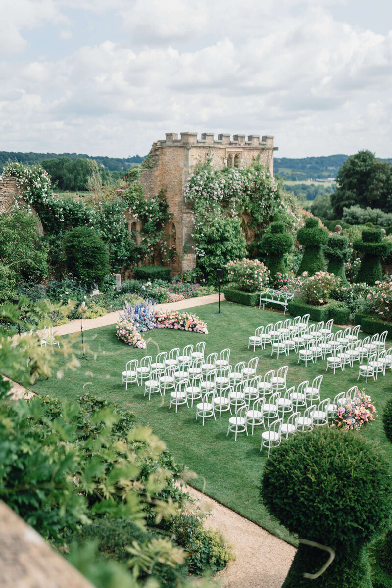 aerial view of a garden wedding ceremony with lines of white chairs and a curved purple and pink floral wall at the top of the aisle