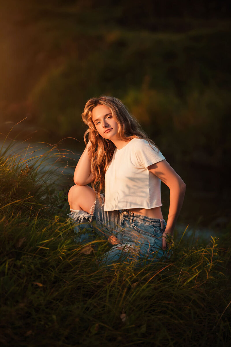 A young woman sitting in grass at sunset, with warm light illuminating her serene expression and flowing hair, captured by a Bemidiji, Minnesota fine art senior photographer