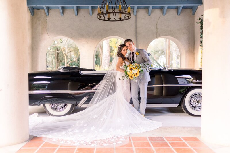Bride and groom stand in front of vintage black car with sunflowers
