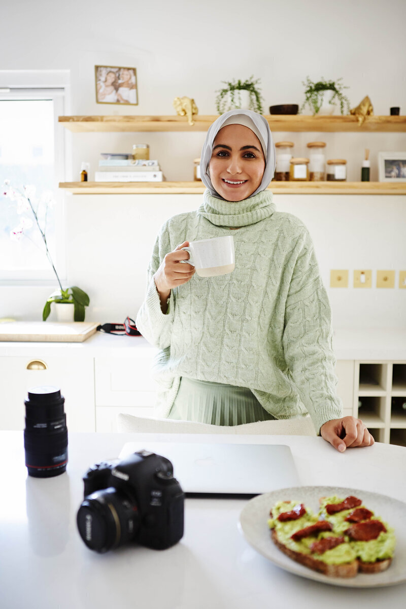 Woman holding a coffee mug while standing in front of a table with a laptop, camera, and plate of food on it.