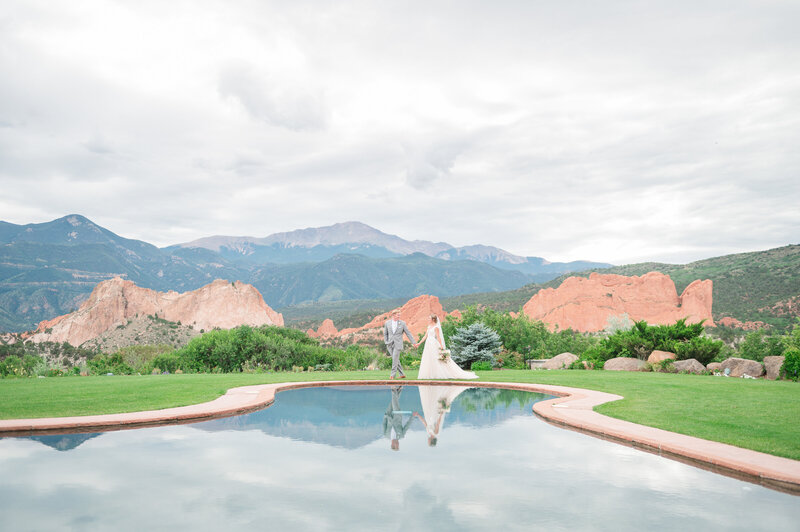 Wide shot of groom leading bride across a lawn in front of an epic mountain background with a reflection pool in front of them.