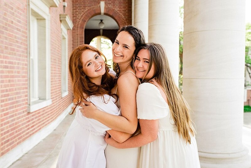 3 Vanderbilt senior girls all wearing white dresses hugging one another and smiling at the camera