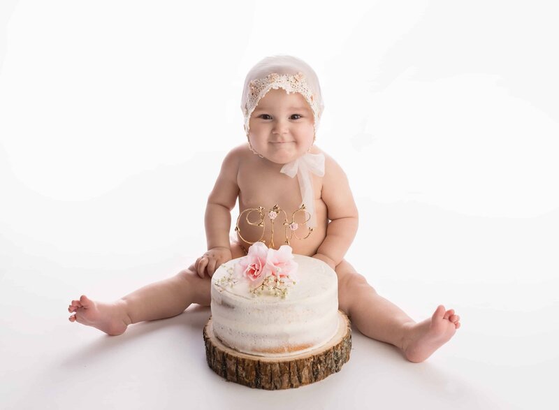 A toddler baby sits smiling behind a cake in a white bonnet on the floor of a Lafayette Baby Milestone Photographer studio