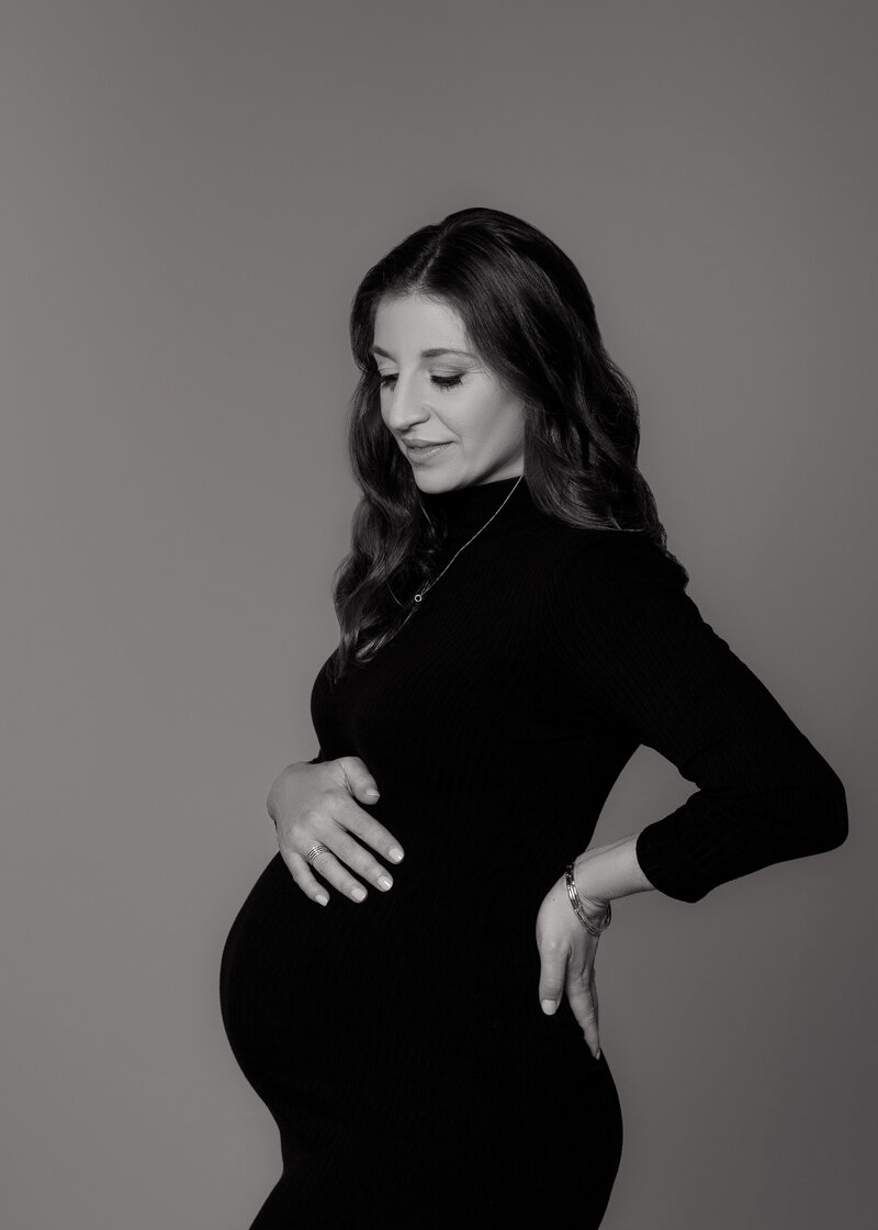 charlotte maternity photographer  for this  mom to be wearing a black long sleeved dress, on a grey background, mom to be is light skinned with dark wavy hair holding her belly and looking down smiling