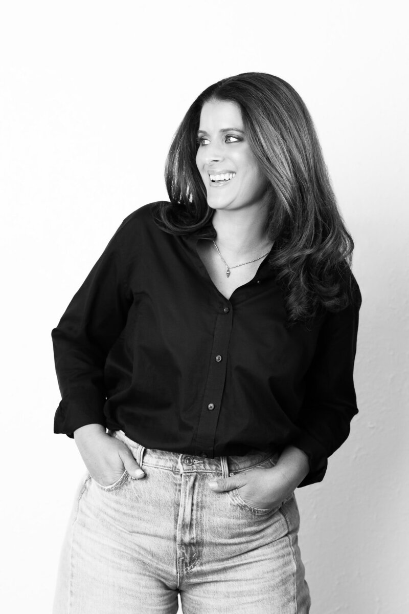 Black & White photo. Woman in black shirt and jeans looking away from camera against white wall.