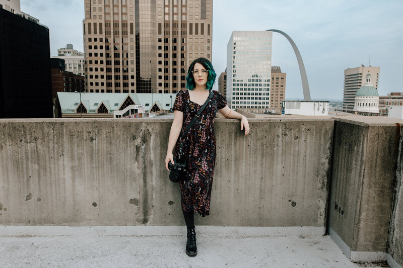 Heather Bennett, owner of Bee Brook Photography, leans against a concrete wall with her camera in her right hand on the rooftop of the Kiener West Parking Garage in St. Louis. The St. Louis Gateway Arch and skyscrapers can be seen in the background.