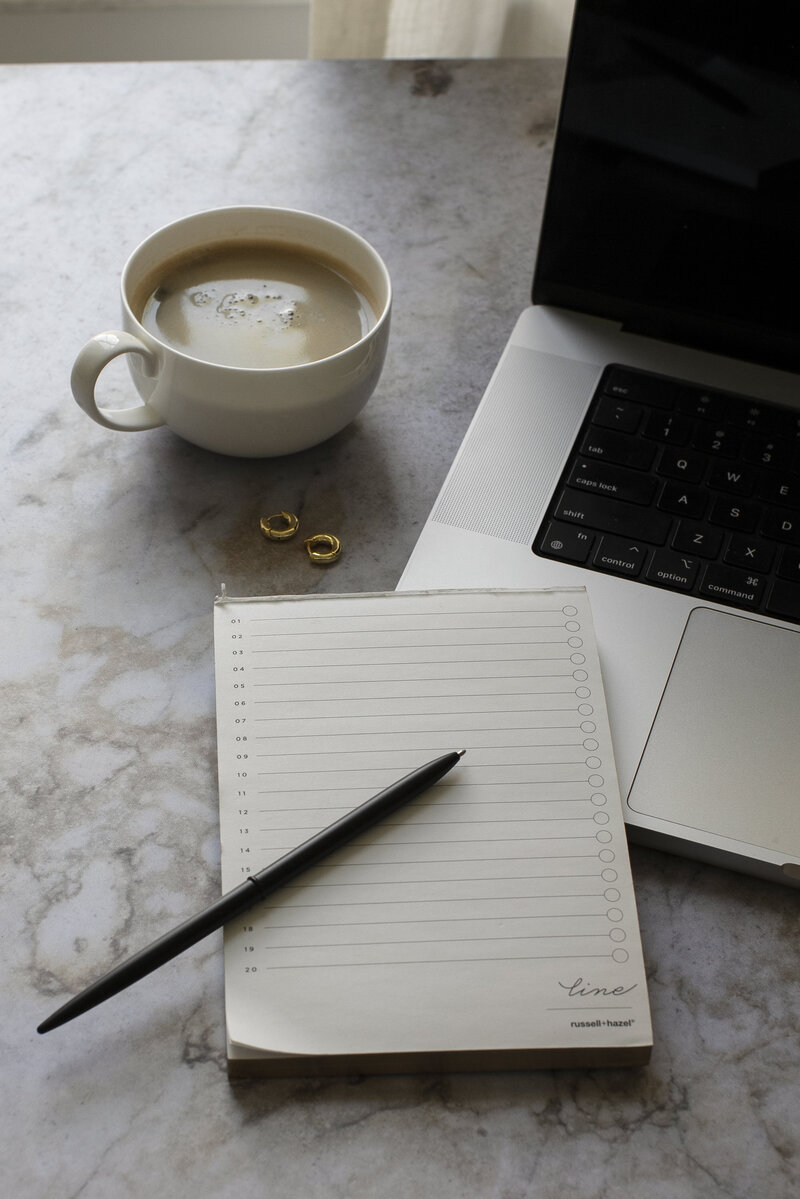 Elegant workspace with coffee cup, small gold hoop earrings, lined notebook with pen, and laptop on marble surface representing thoughtful reflection and planning.
