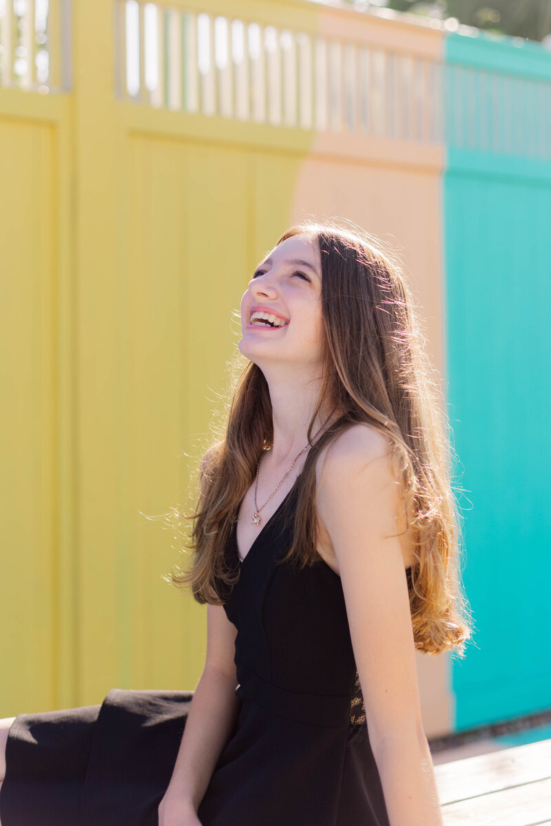 A young girl laughing up at the sky in a black dress in front of a yellow and green fence in Galveston Texas
