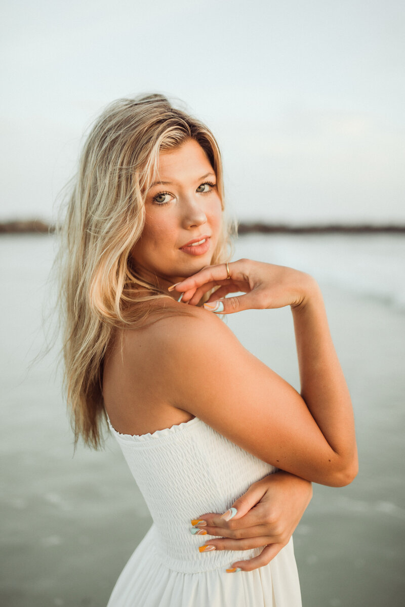 young blonde senior poses in white dress on beach in st. andrews state park. Portrait by Brittney S of Be Seen Photos