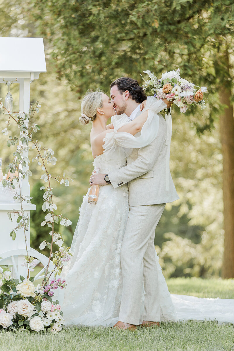 Bride and Groom share a kiss at their french countryside wedding. They are smiling. Bride's arms are draped around groom's neck and holding a bottle of champagne. Captured by top destination wedding photographer Lia Rose Weddings.