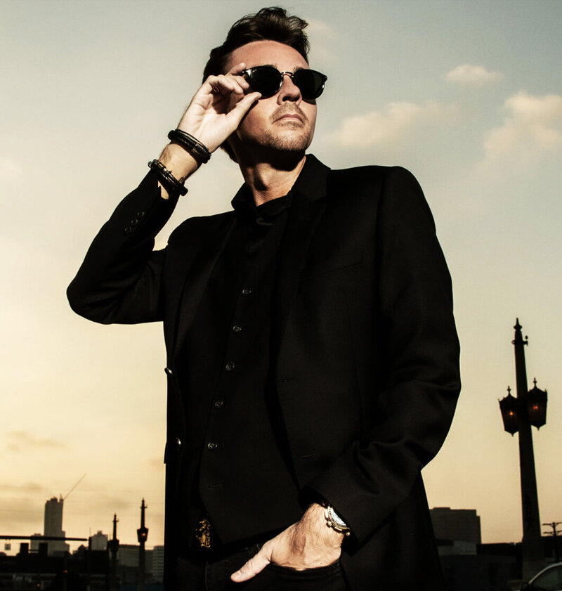 Author Branding Portrait Chandler Morrison wearing black suit hand to sunglasses he wears standing on street against sun setting behind him