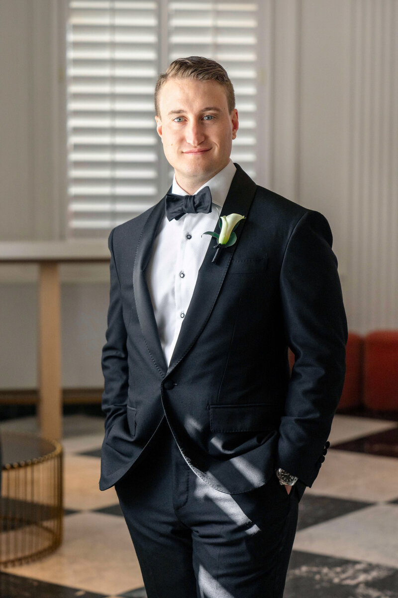 A person wearing a black tuxedo with a white shirt and bow tie stands indoors. The room has light-colored walls and a floor with black and white geometric patterns. They are smiling with hands in pockets.