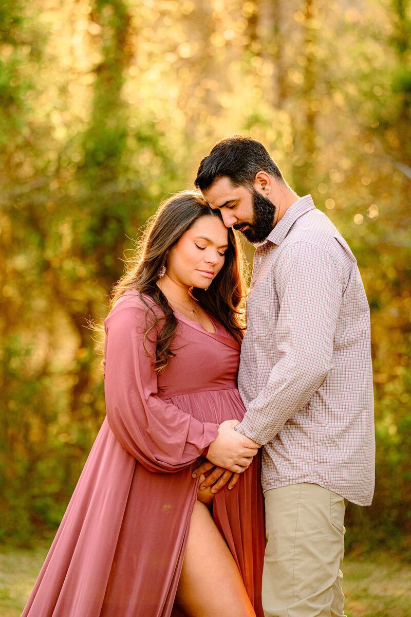 And expectant couple has both of their heads brought together mom is dressed in a rose pink dress dad and a button-down shirt both with their hands on mom's belly