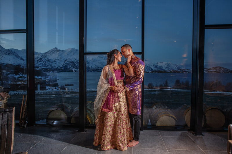 Inside a room with a vast glass facade offering a view of a snowy Norwegian landscape at dusk, a bride and groom stand close together, creating a picturesque moment. The bride is adorned in a traditional South Asian lehenga, rich in gold and pink embroidery, and the groom wears a matching purple sherwani with a golden dupatta. They look into each other's eyes with affection, framed by the natural splendor of the fjord and mountains in the glass behind them. The warmth of their embrace contrasts with the cool tones of the evening outside, capturing the intimate celebration of their wedding day.