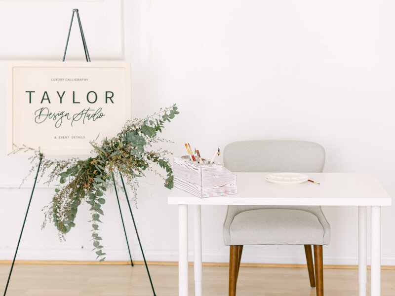 A sign bearing the name 'Taylor Design Studios' placed beside a table adorned with various calligraphy supplies