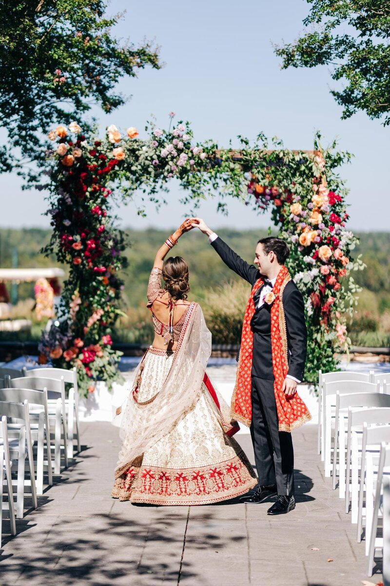 A couple dressed in traditional Indian wedding attire dancing under a floral archway at an outdoor ceremony, captured by a luxury wedding photographer.