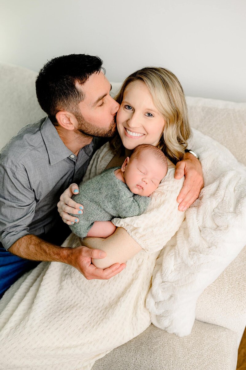 father kisses his wife's temple as their newborn snuggles on hier shoulder
