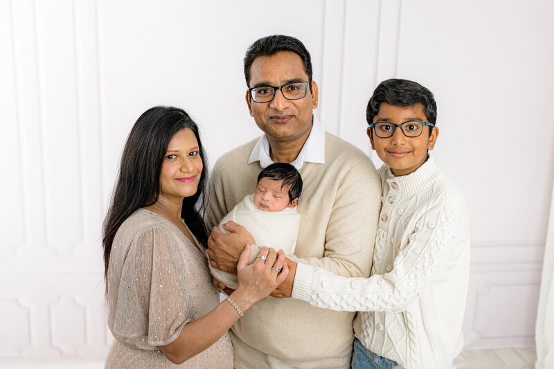 Father holds his newborn son while the entire family is dressed and neutral tones with mother on his right hand side and his oldest son behind him each with their hands on the new baby