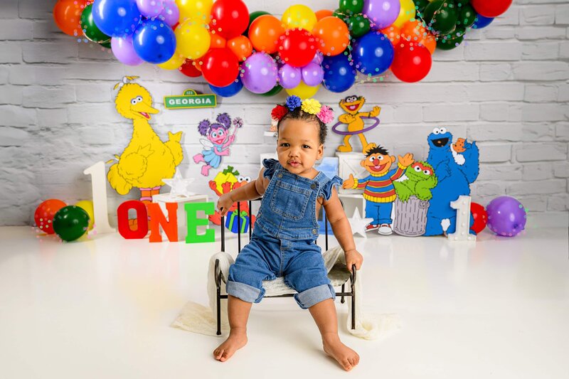 little girl sits on a bench dressed in overalls with a sesame street background