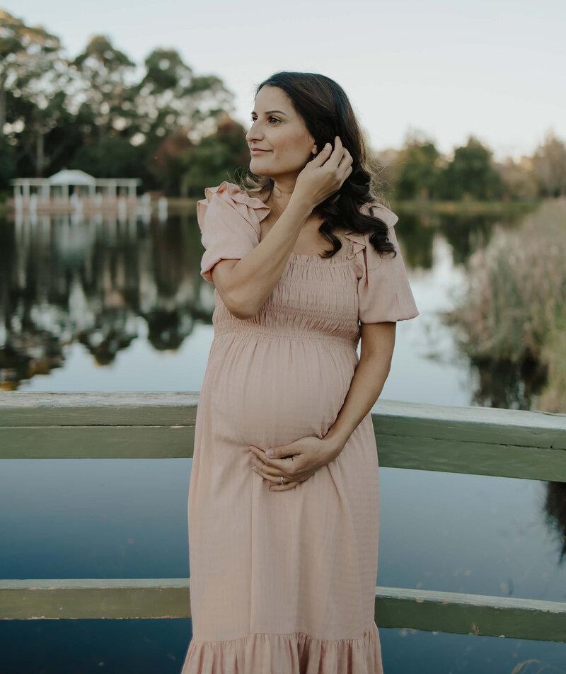Pretty pregnant woman in pink dress posing on a green bridge over a pond in a garden while holding her belly and pushing her hair back gently behind her ear
