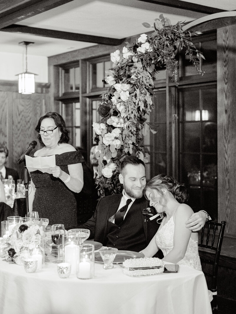 Black and white photo of mom giving speech at wedding reception while bride and groom hug