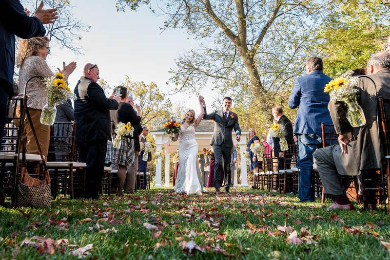 A bride and groom celebrate after their wedding ceremony, holding up their hands while guests clap