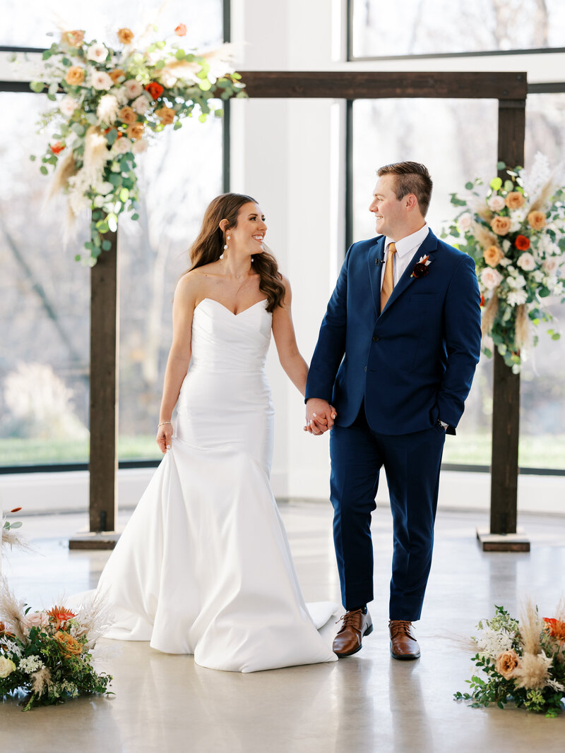 bride and groom holds hands in Osage House Chapel walking towards camera looking at each other with arbor behind them decorated with fall florals